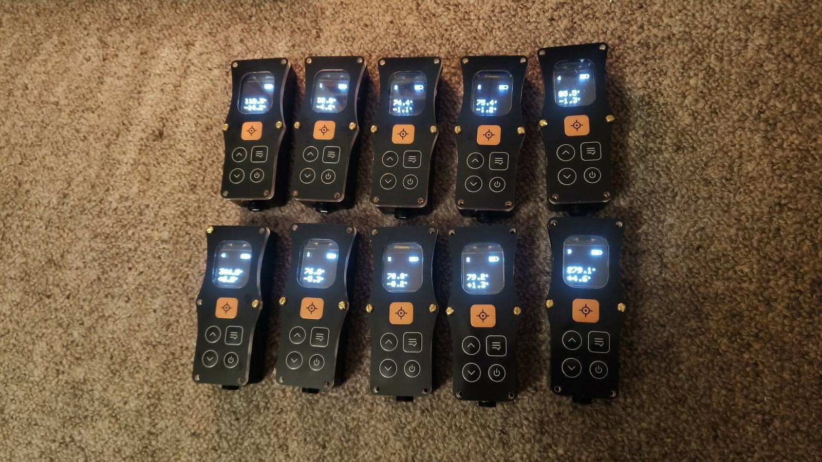 Ten DiscoX v2 devices laid out in two rows of five on a beige carpet, all switched on. Each has a black case with brass screws around the edge, a lit screen at the top showing a bearing and an inclination reading, a square orange trigger button with a crosshair icon below the screen, and four round buttons — up, menu, down and power — beneath that. A black connector gland sits at the bottom end of each case.