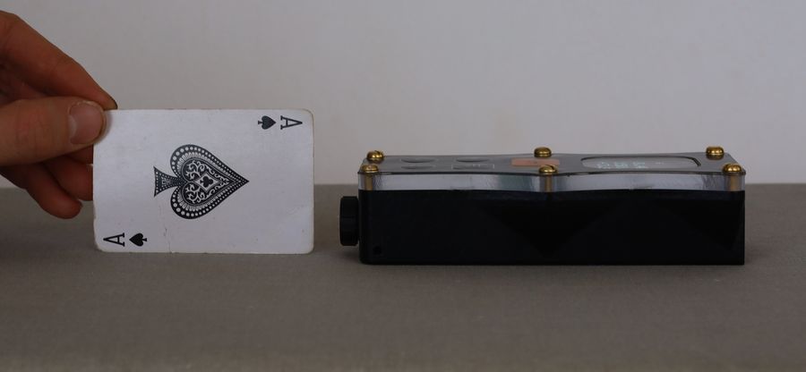The DiscoX lying on its side next to a playing card held upright, showing the clear top plate, the brass screws along its length and the lens housing at one end.